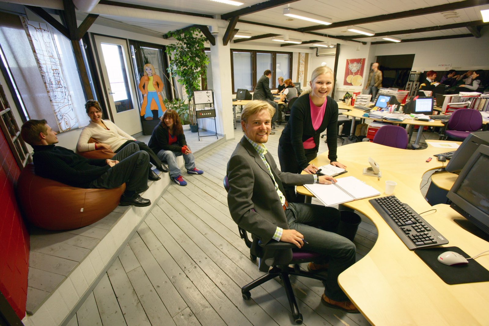 Open-plan office workspace with people collaborating at desks and an exposed structural ceiling overhead